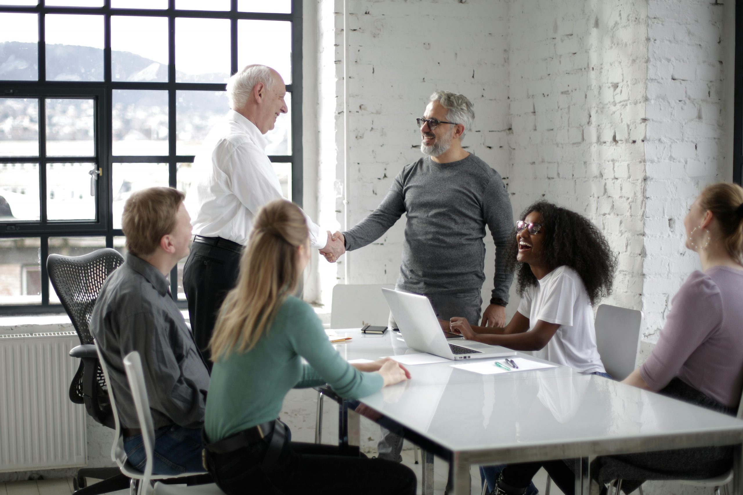 Cheerful male colleagues shaking hands while discussing business ideas with group of multiethnic coworkers gathering around table with gadgets and documents in modern light workspace